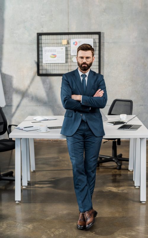 full length view of confident bearded businessman in suit and eyeglasses standing with crossed arms