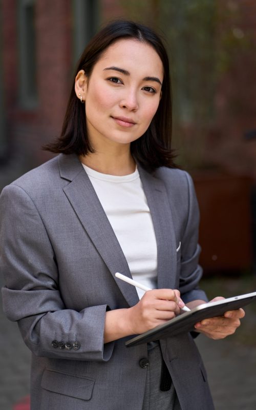 Young confident elegant professional leader Asian business woman, female executive manager consultant, entrepreneur wearing suit holding tablet standing outside office looking at camera, vertical.
